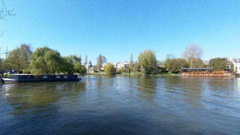 A pretty view of a river with canal boats and trees in the foreground. 