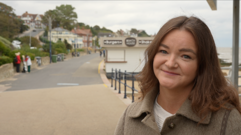 A head and shoulders image of Jenny Riddell-Carpenter, who is standing on the promenade in Felixstowe, smiling and looking into the camera. There is the beach sea behind her to the right and to left you can see houses going up a road. 