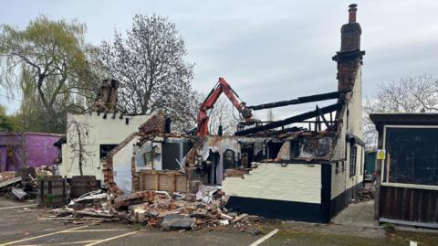An old pub which is in the process of being demolished. There is a digger behind it. Only sections of the outer wall remain and there are bricks on the ground. The toilet is visible in the front.