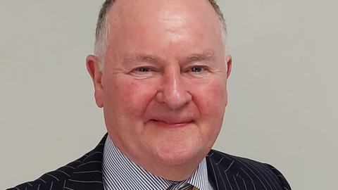 A head and shoulders shot of a man in a pin striped suit, with a dark blue striped shirt smiling at the camera