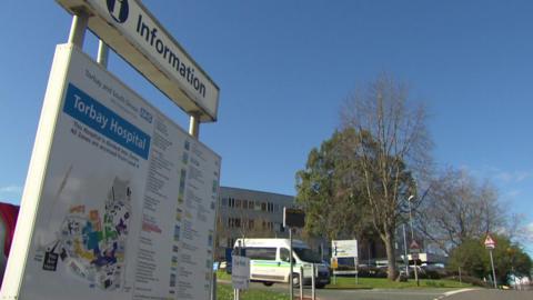 A Torbay Hospital sign is displayed on the site. The sky is bright blue. Trues and road signs are in front of a hospital building.