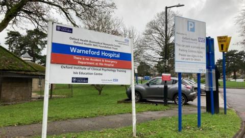 A sign outside a hospital saying Warneford Hospital. There's a carpark in the background and some autumnal trees.