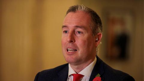 Paul Givan speaking. He is wearing a black suit jacket, white shirt and red tie.