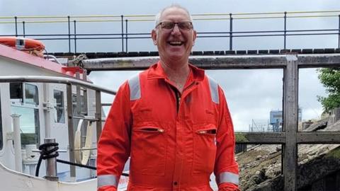 Tony Cowell, in a bright orange jumpsuit, beams at the camera as he stands beside the ferry. He has short white hair and brown-rimmed glasses.