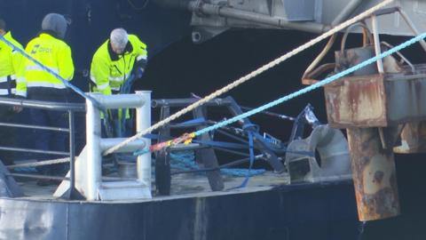Three people in yellow high-vis jackets with DFDS branding on them inspect the damage caused to the ferry firm's Tarifa Jet vessel in St Helier, Jersey, after it hit a berth in the harbour. Railings have been bent out of shape. White and blue ropes have been tied to the boat.