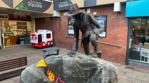 A statue of a man wearing traditional pilgrim clothing kneels on top of rock on St Andrew's Square in Droitwich. The year 1620 is carved into the rock.