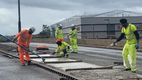 Workers in yellow and orange high-vis outfits are working on a road laying tarmac. It is a cloudy day.