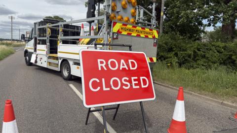 A road has been closed and has cones going across it. There is a sign in the middle of the road which is red and has ROAD CLOSED on it in white writing. A truck is parked across the road.