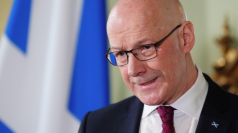 John Swinney in dark suit and white shirt with a burgundy tie in front of a Saltire flag