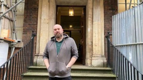 A man with a shaved head and light-brown beard stands in front of the grand stone entrance to a red-brick Georgian house. He is wearing a grey hoody and green polo shirt and has his hands in his pockets. Scaffolding can be seen to either side of the doorway.