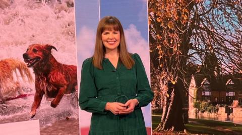 Alexis Green stands in the 91ȱ South studio in front of a screen showing the pictures of the day, which include one of a golden retriever dog running in the waves on the beach and a tree in a village courtyard. Alexis smiles at the camera, with long brown hair and wearing a forest green long sleeved dress