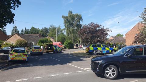 Four police cars at the entrance to a residential road. A black car is turning into the road. Officers can be seen clustered outside a red brick house in the background.