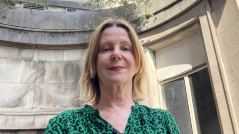Maria Kane pictured standing outside of an old building at the Bristol Royal Infirmary. She has short blonde hair and is wearing a green leopard print dress. She is looking down at the camera