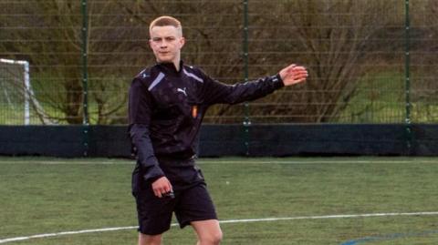RJ refereeing a football game. He stands on a football pitch his left arm extended. He is wearing a black sports jacket and black shorts.