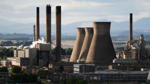 An oil refinery with tall buildings emitting smoke. Mountains are seen in the background.