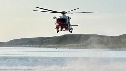 A coastguard helicopter hovers over the sea. There are a handful of houses below a cliff in the background