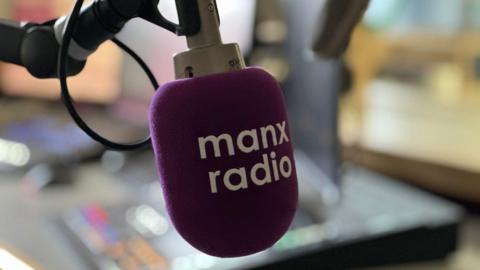 A microphone in a radio studio which has a purple pop shield over it with Manx Radio in white lettering across its face.