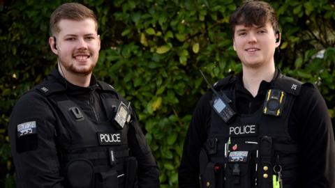 PCs Owain Soady-Jackson and Morgan Llewellyn. They are both wearing black police uniform and have short dark hair. Behind them is a bush.