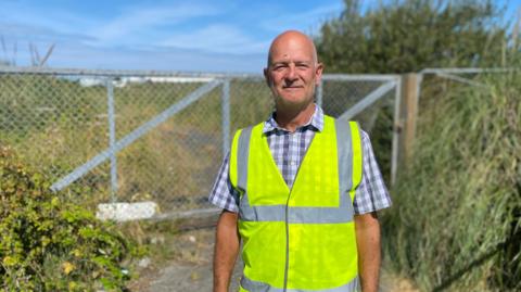 A man wearing a hi-vis jacket and a checked shirt, standing in front of a wire fence blocking an overgrown field.