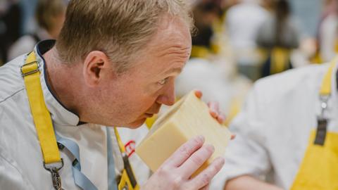 A person in a yellow apron smells a big chunk of cheese