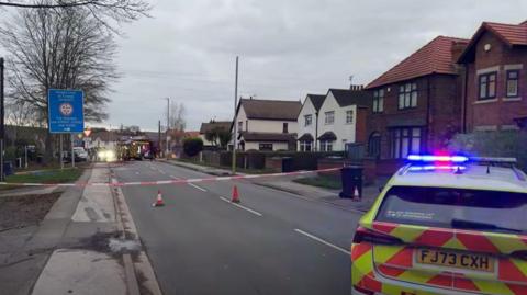 Still from fire service footage showing a residential road which has been sealed off with warning tape and cones. In the foreground is a police car with a fire appliance visible further along the road