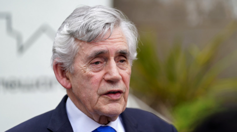 Headshot of former Prime Minister Gordon Brown, wearing a  navy suit and white shirt and tie.