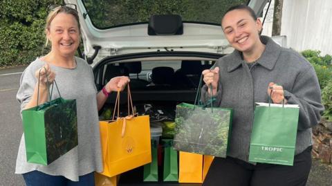 Two women stand smiling around the back of a white car boot. They are carrying bags containing Christmas gifts. The bags are green and yellow.