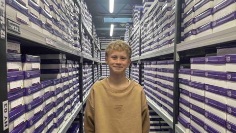 A blond boy wearing a brown sweatshirt is stood in a warehouse setting with boxes on either side of him.