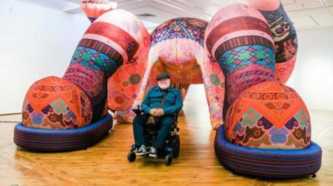 Mr Wilsher-Mills sits in a powered wheelchair underneath a giant blow up colourful sculpture of what appears from the camera angle to be a person lying down with their feet on the floor and knees bent.