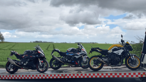 Three motorbikes, black with red, blue and yellow features attached to a trailer which has red and white checked sides. A farmer's field can be seen behind them.