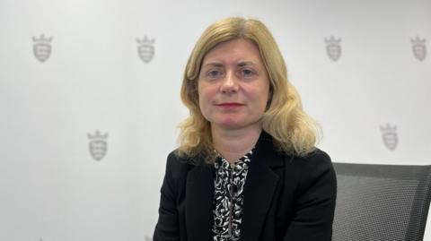 Deputy Lyndsay Feltham is sitting down and faintly smiling at the camera. She has shoulder-length blonde hair and blue eyes. She is wearing a black jacket over a black and white patterned blouse. Behind her the wallpaper features the Jersey shield of arms design.