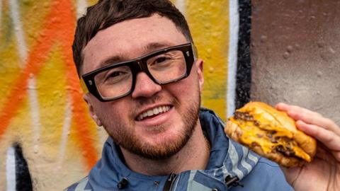 Oscar is wearing dark rimed glasses and is smiling at the camera whilst holding up a cheeseburger. He's standing in front of orange graffiti.