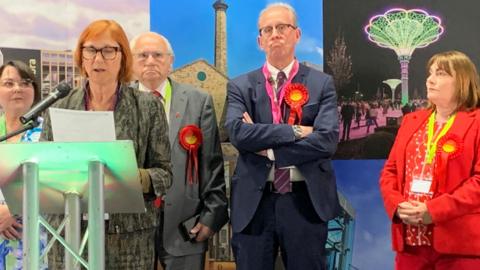 An election count, with four people standing behind a woman reading out results from a piece of paper. Two men and a woman all wearing red rosettes look disappointed.