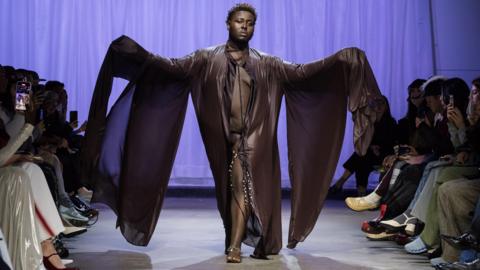 Model James Corbin at London Fashion week. He is wearing a brown robe, walking down the catwalk with his arms out wide.