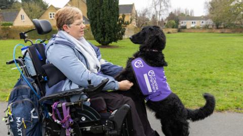 Samantha, a woman, with short golden hair. She is sitting on a motorised wheelchair with her black dog standing on it's two legs looking at her face. 