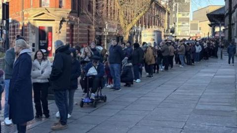 A queue of people down a street in Preston. Many are wearing coats and bracing from the cold. 