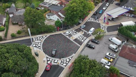 An aerial photo of two roundabouts, with rainbow leaves painted on the tarmac. At each of the the three exists for each roundabout there's a band of white leaves painted across the road.
