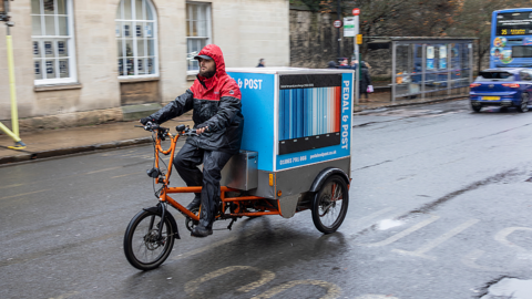 Delivery Cyclist Pedaling An Orange Cargo Bike With Mailbox On Rainy City Street.
