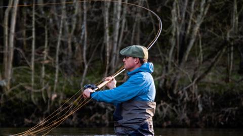 A man with a green cap, blue waterproof top and black waders stands up to his waist in water while casting a fishing rod
