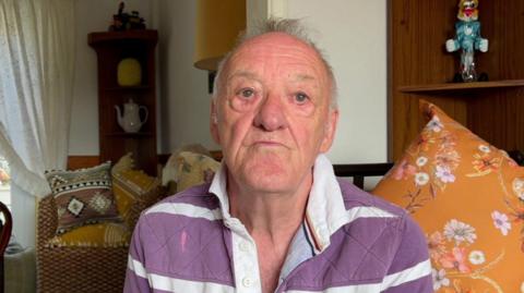 An elderly man looking straight at the camera. He has a stern look on his face and is wearing a purple and white striped shirt. He is sat on a chair in his living room with lots of cushions and ornaments behind him. 