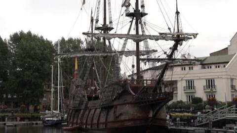 A brown17th century large ship with sails in the water near some white buildings.