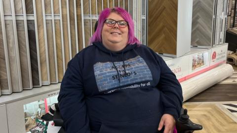 Liz Owens wearing a navy blue hoodie with access in St Neots written in the centre. She has pink hair, black and pink glasses and is smiling at the camera. Liz is in her wheelchair at the Floor by Ian shop. Behind her are various laminate and wooden floor designs. 