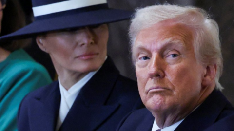 US President Donald Trump looks down the lens on the inauguration day of his second Presidential term with first lady Melania behind him, wearing a large navy hat.