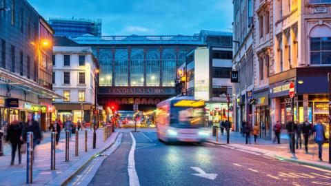 A photo taken of Glasgow Central Station and Argyle street in the evening with illuminated shop signs and the pavements busy with pedestrians