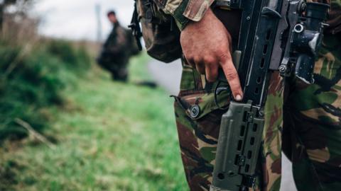 A soldier holding a gun. They are stood on the side of a road on an area of grass.