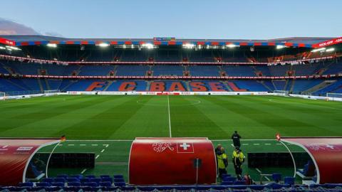View of St Jakob-Park stadium taken from behind a red dugout on the halfway line, with the name FC Basel in red text on a blue background on the seats opposite