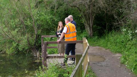 Two men and a woman stand at a jetty overlooking a pond. They are surrounded by greenery. 