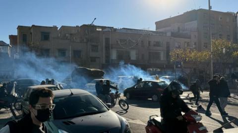 A street-view image of Iranian shopkeepers and traders protesting on motorbikes and on foot in between cars with tear gas is visible.