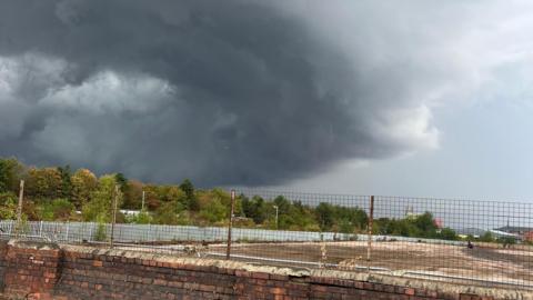 A large grey shelf cloud moving over a row of trees and a river 
