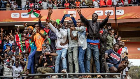 A large group of male Kenya fans pack on to a raised section obviously not designed for them at the Kasarani Stadium, with many clinging on to gain a vantage point. More fans can be seen in the stand behind them, which has orange CHAN branding and sponsors logos on it